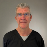 Portrait of a middle-aged man with gray hair and glasses, wearing black scrubs, against a neutral gray background.
