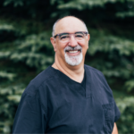 Smiling middle-aged man in navy medical scrubs outdoors, greenery in the background.