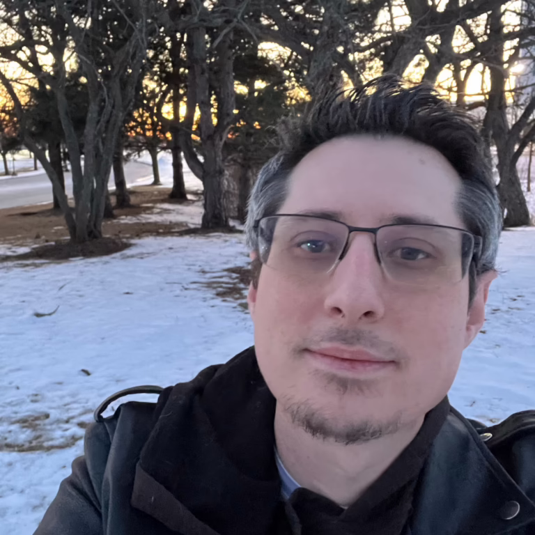 Close-up selfie of a man with glasses in a snow-covered park at sunset light filtering through bare trees.