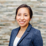 Smiling Asian woman in a blue textured blazer posing against a neutral stone wall background