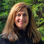 Woman with long brown hair smiling outdoors, wearing a black jacket, in front of green foliage in sunlight.