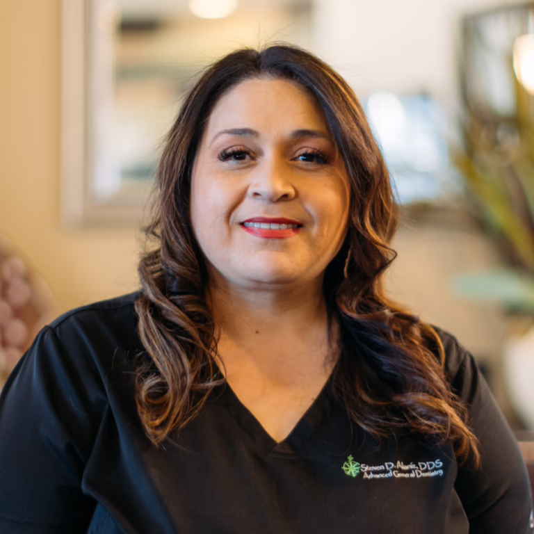 Portrait of a smiling woman in black dental scrubs with a dentistry logo on the chest, in a clinic setting.