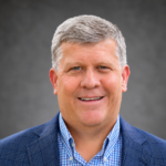 Professional headshot of a smiling man with gray hair, wearing a blue blazer and checkered shirt against a neutral background.