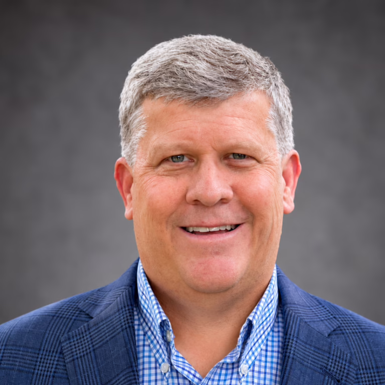 Professional headshot of a smiling man with gray hair, wearing a blue blazer and checkered shirt against a neutral background.