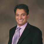 Smiling man in a black suit with a lavender shirt and matching tie, posing for a professional portrait against a neutral backdrop.