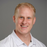 Head-and-shoulders portrait of a smiling man with blond hair in a white polo against a gray backdrop.