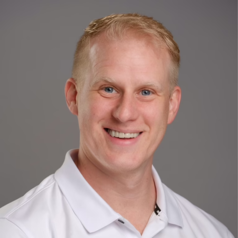 Head-and-shoulders portrait of a smiling man with blond hair in a white polo against a gray backdrop.