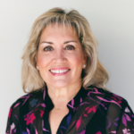 Portrait of a smiling woman with blonde hair wearing a dark floral blouse against a light background.
