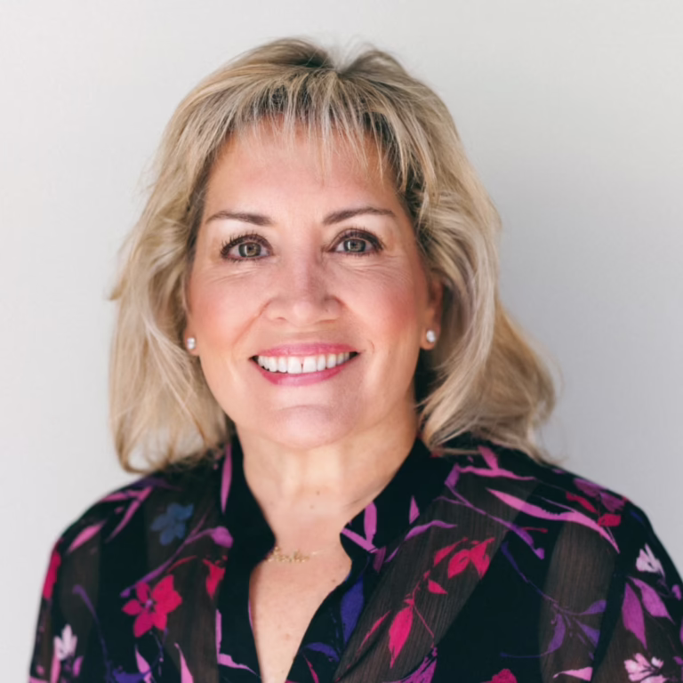 Portrait of a smiling woman with blonde hair wearing a dark floral blouse against a light background.