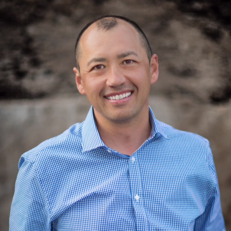 Smiling man in a blue checkered shirt outdoors, posing for a professional headshot.