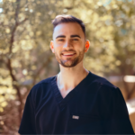 Portrait of a smiling man in navy scrubs outdoors, with a blurred natural background behind him.