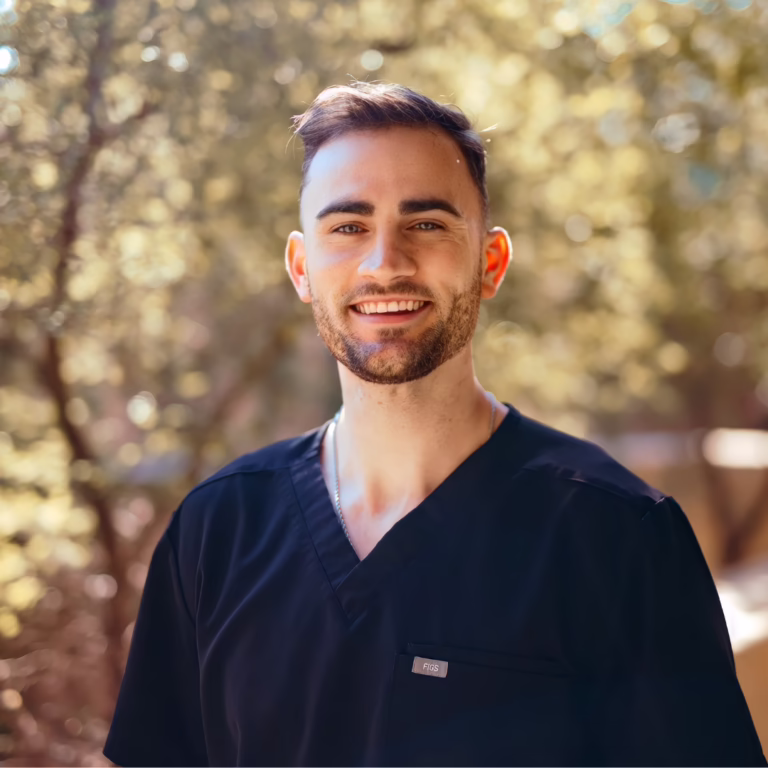 Portrait of a smiling man in navy scrubs outdoors, with a blurred natural background behind him.