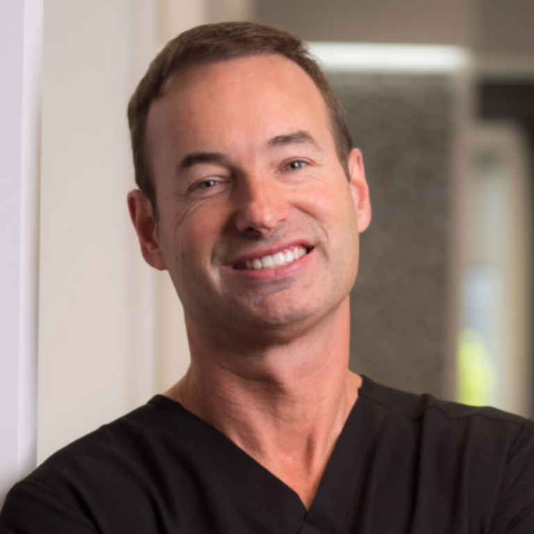 Smiling man in black scrubs standing in a bright clinical hallway.