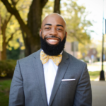 Smiling man in a gray suit and yellow bow tie posing outdoors, sunny park background.