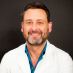 Smiling male doctor in a white coat and blue shirt against a dark background.