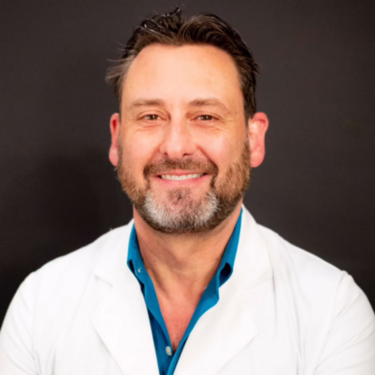 Smiling male doctor in a white coat and blue shirt against a dark background.