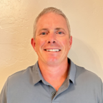 Smiling man with short gray hair wearing a gray polo shirt, standing against a beige textured wall.