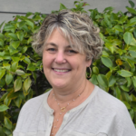 Smiling woman with short, wavy gray-blonde hair and hoop earrings, standing in front of green shrubs; wearing a light gray blouse and a delicate necklace.