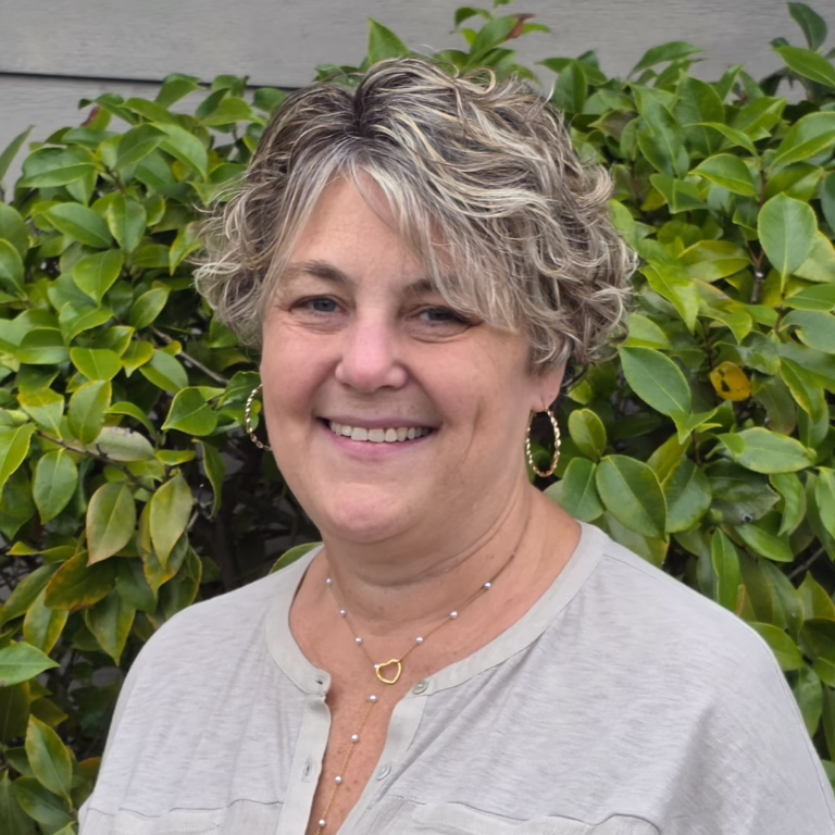 Smiling woman with short, wavy gray-blonde hair and hoop earrings, standing in front of green shrubs; wearing a light gray blouse and a delicate necklace.