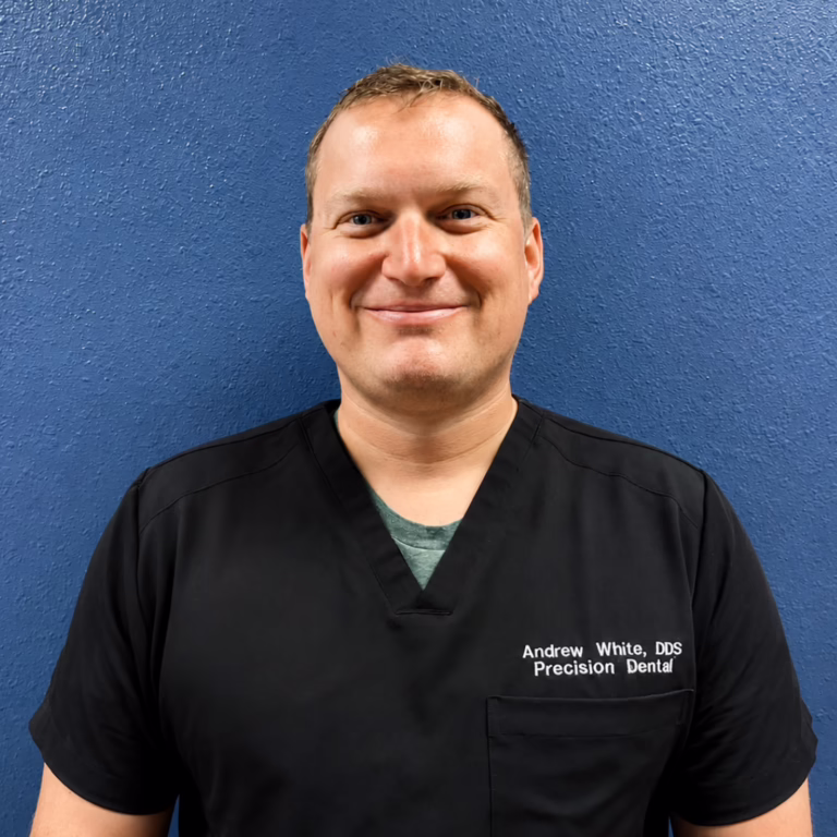 Smiling man in black dental scrubs against a blue wall; name reads Andrew White, DDS, Precision Dental.