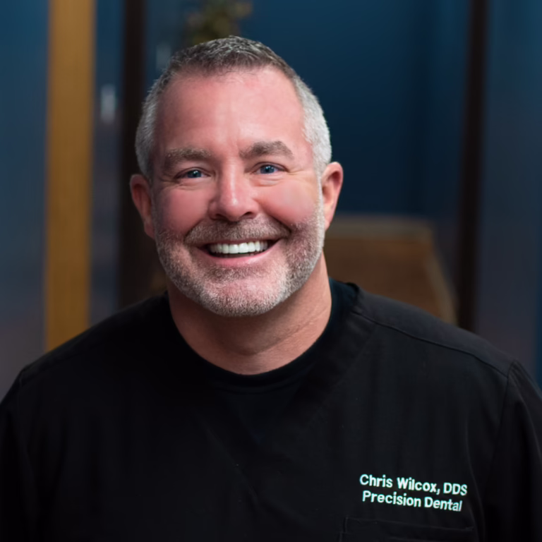 Portrait of Dr. Chris Wilcox, DDS, smiling in black scrubs with his name and clinic embroidered on the chest: Precision Dental.