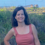 Smiling woman with shoulder-length brown hair in a pink sleeveless top, standing among tall coastal grasses with a wooden boardwalk behind her.