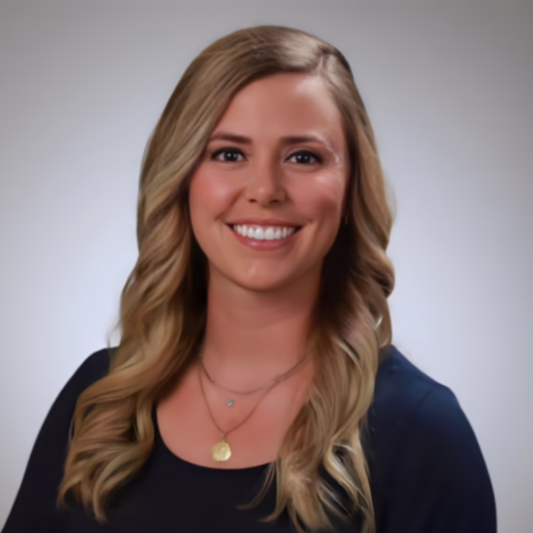 Portrait of a smiling woman with wavy blonde hair, wearing a dark top and layered necklaces against a gray gradient background.
