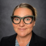 Head-and-shoulders portrait of a woman wearing glittery cat-eye glasses and a black blazer against a gray studio backdrop. great