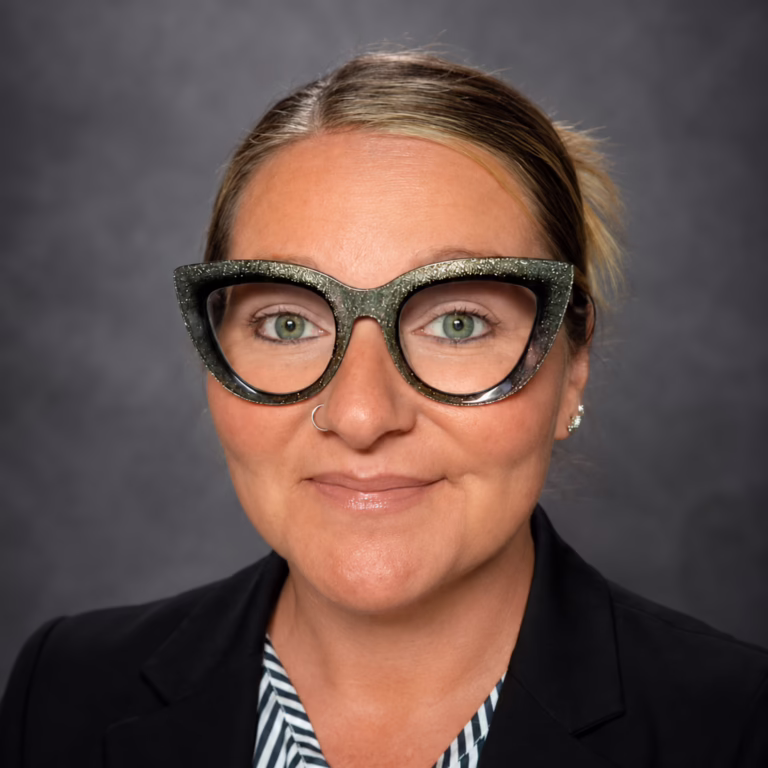 Head-and-shoulders portrait of a woman wearing glittery cat-eye glasses and a black blazer against a gray studio backdrop. great