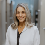 Smiling female clinician in a white lab coat standing in a clinic hallway.