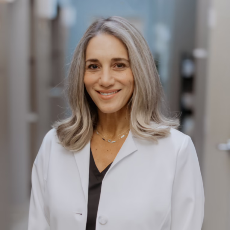 Smiling female clinician in a white lab coat standing in a clinic hallway.