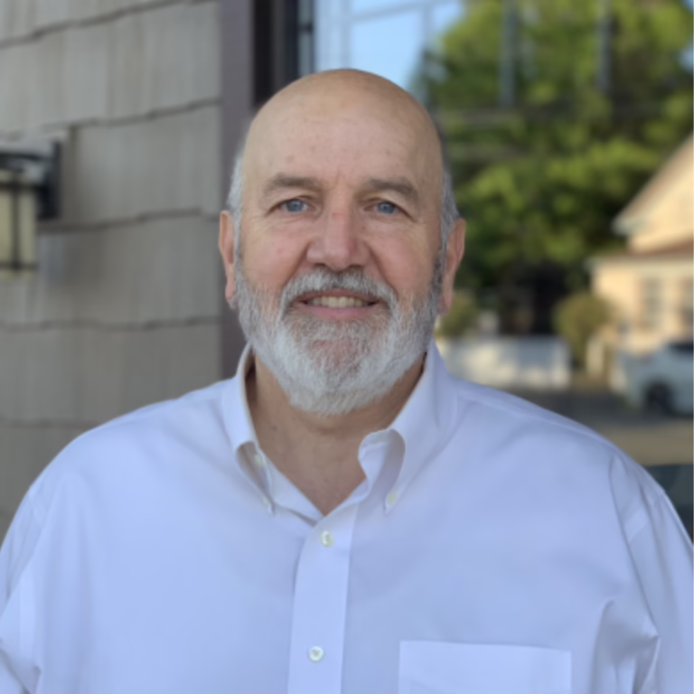 Smiling older man with a white beard wearing a white button-down shirt outdoors near a building.