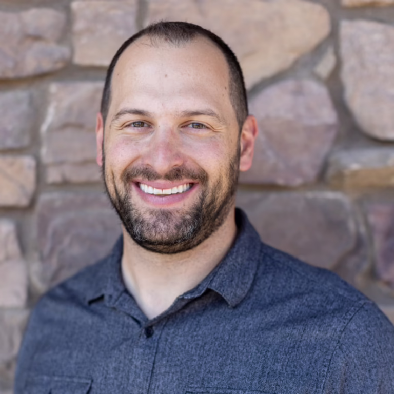 Smiling man in a dark blue shirt poses against a stone wall backdrop (head-and-shoulders portrait).
