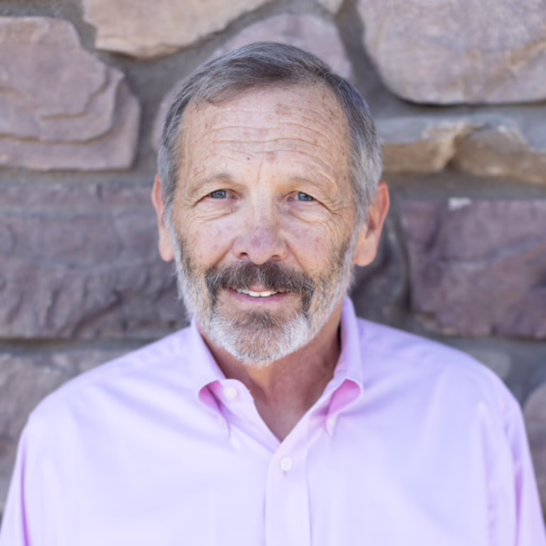 Smiling older man with gray hair and beard in a pink button-down shirt, standing against a stone wall background.