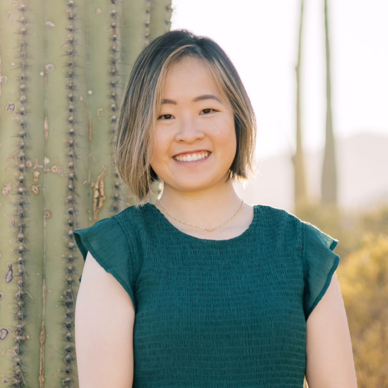 Smiling young woman with short, light-brown hair standing beside a cactus outdoors in warm sunlight, wearing a dark green textured top.
