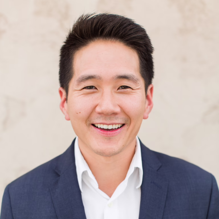 Smiling man in a navy blazer and white shirt posing against a neutral background (professional headshot)